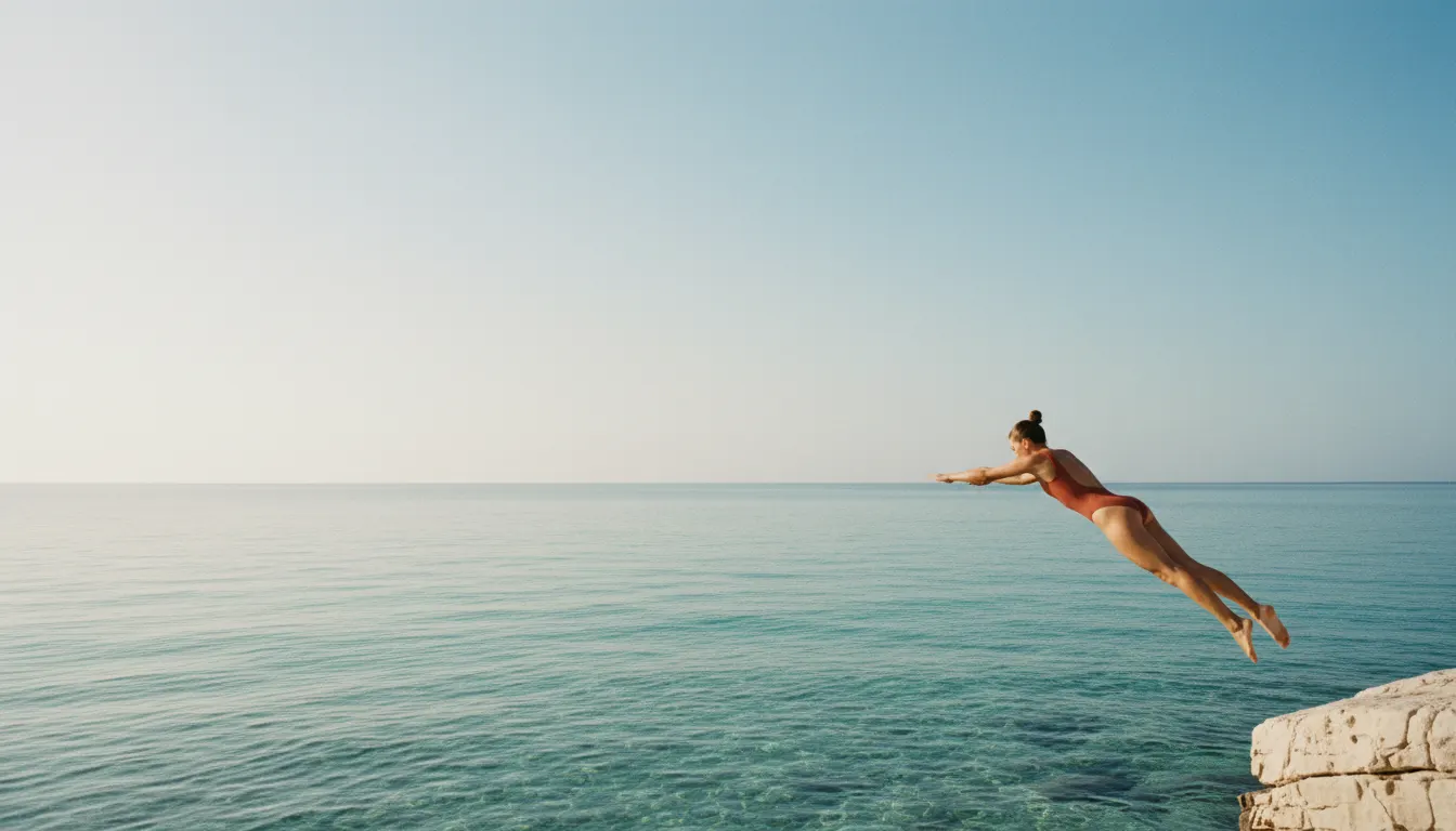 A swimmer in the terracotta Marina scoop-back one-piece diving off a pale limestone rock into clear turquoise Mediterranean water at mid-morning, caught mid-air in the lower right of the frame, with the upper left half of the picture left as open, cloudless pale-blue sky softened by sun haze